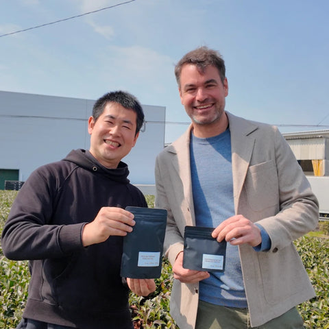 Oishi farmer and Nio Teas' CEO standing outdoors in a green tea plantation holding green tea pouches with white labels under a blue sky.