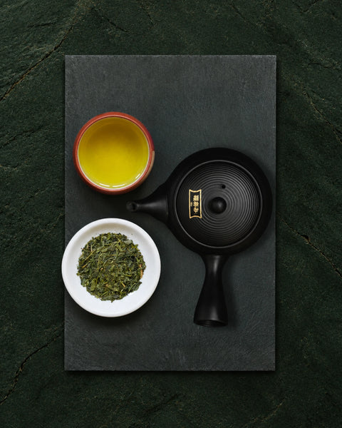 A top-down flat lay of a black teapot with gold detailing, a small white bowl of bancha loose green tea leaves, and a small red-rimmed cup filled with yellow tea, arranged on a dark slate board against a dark green textured background.