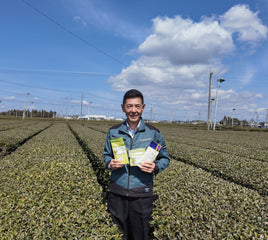 Oishi farmer standing in a tea plantation holding three product pouches, with rows of tea plants, utility poles, and a blue sky with clouds in the background.