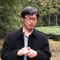 Noike farmer wearing black-rimmed glasses, a black coat over a blue and white checkered shirt, standing outdoors in front of a green hedge and trees.