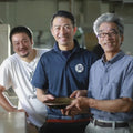 Three generations of Osada farmers standing together in what appears to be a pottery or ceramic workshop, with the central man holding a small round ceramic dish. The man on the left wears a white t-shirt, the center man wears a navy blue polo shirt with a white circular logo, and the man on the right wears a blue-and-white checkered button-up shirt.
