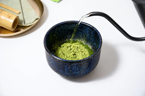 A dark blue speckled ceramic bowl filled with green matcha powder, with hot water being poured into it from a black gooktoe kettle on a white surface.