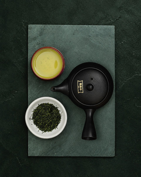 A black ceramic teapot with gold calligraphy on the lid, arranged on a dark green slate tray alongside a small bowl of sencha loose green tea leaves and a cup of yellow tea, against a dark textured background.